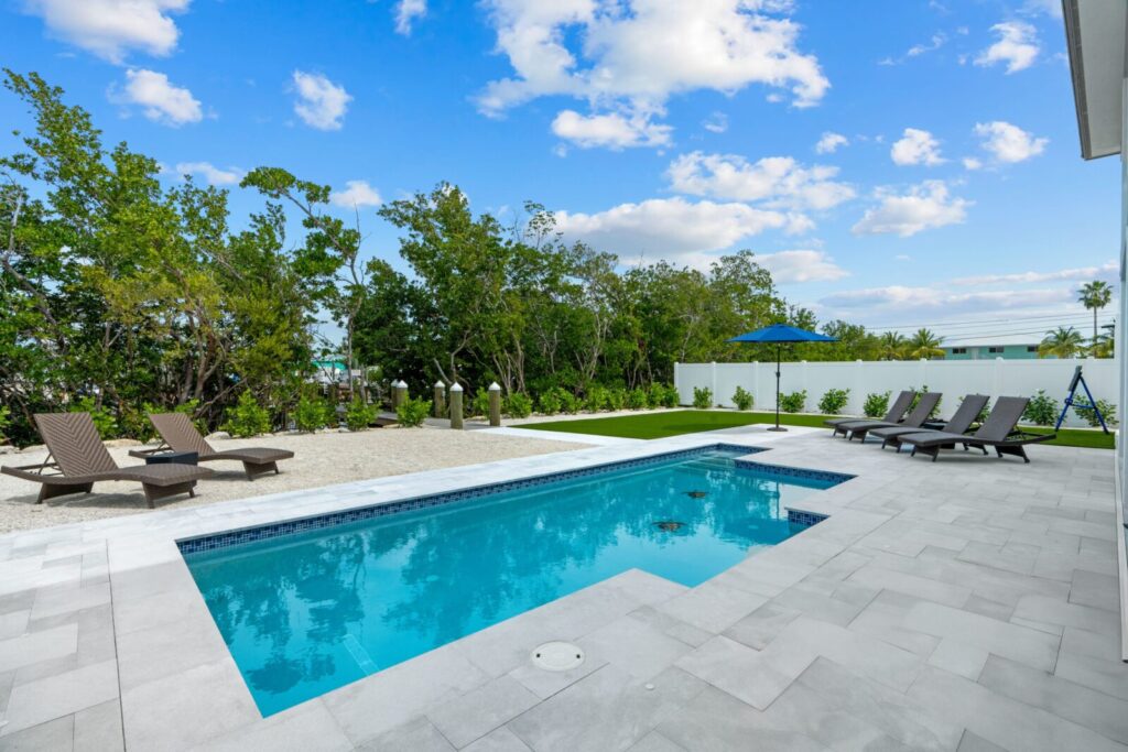 Modern backyard with a rectangular pool, surrounded by light gray patio tiles, several lounge chairs, green lawn, trees, and a blue umbrella under a partly cloudy sky.