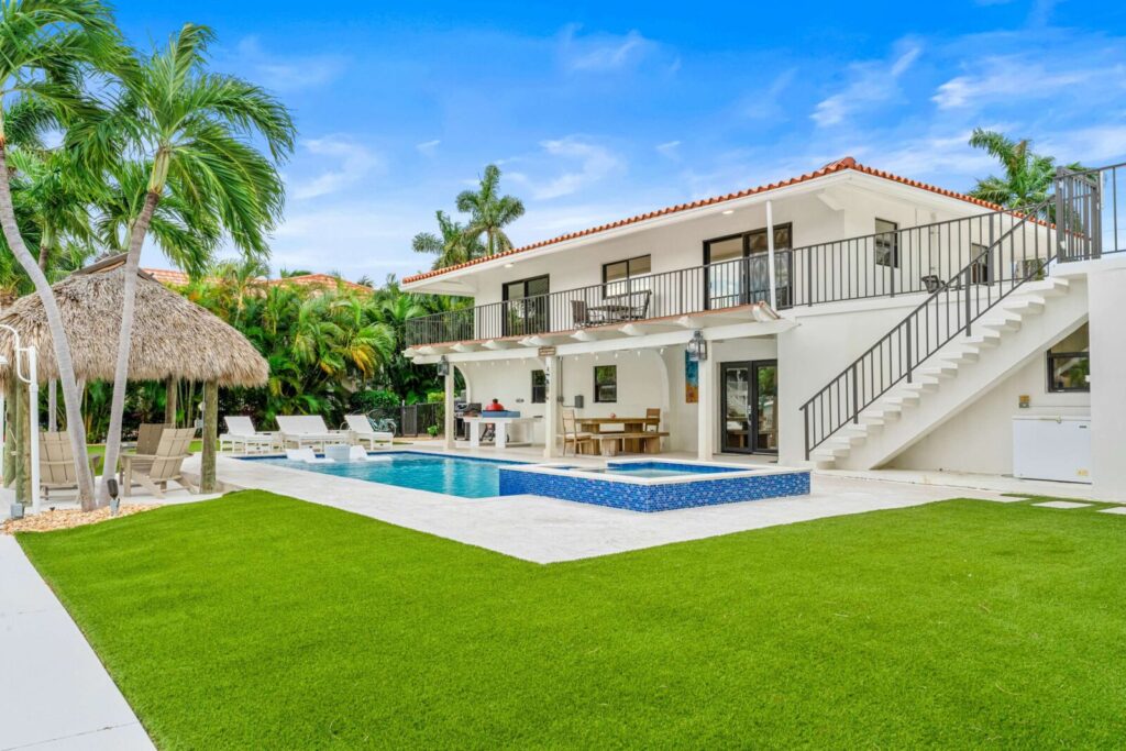 A modern two-story house with a red tile roof, large balcony, and outdoor pool area, surrounded by green lawn, palm trees, lounge chairs, and a thatched pergola, under a bright blue sky.