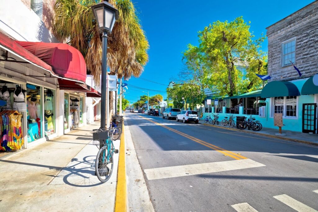 A sunny street lined with shops, palm trees, and parked bikes, featuring colorful awnings and a clear blue sky in a relaxed, tropical town setting.
