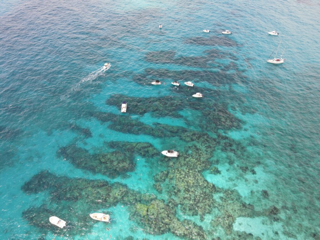 Aerial view of clear turquoise water with patches of reef visible below the surface. Several small boats are floating and moving across the water, scattered throughout the scene.