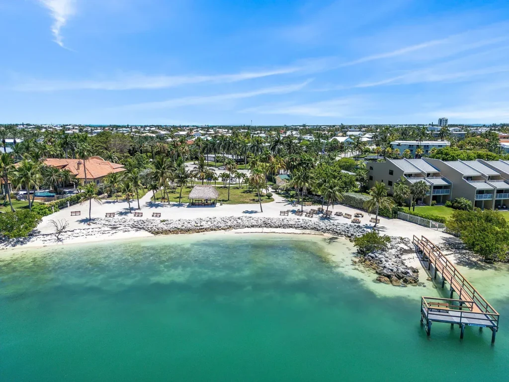 Aerial view of a tropical beach with white sand, clear turquoise water, palm trees, lounge chairs, a pier, and buildings surrounded by greenery under a bright blue sky.