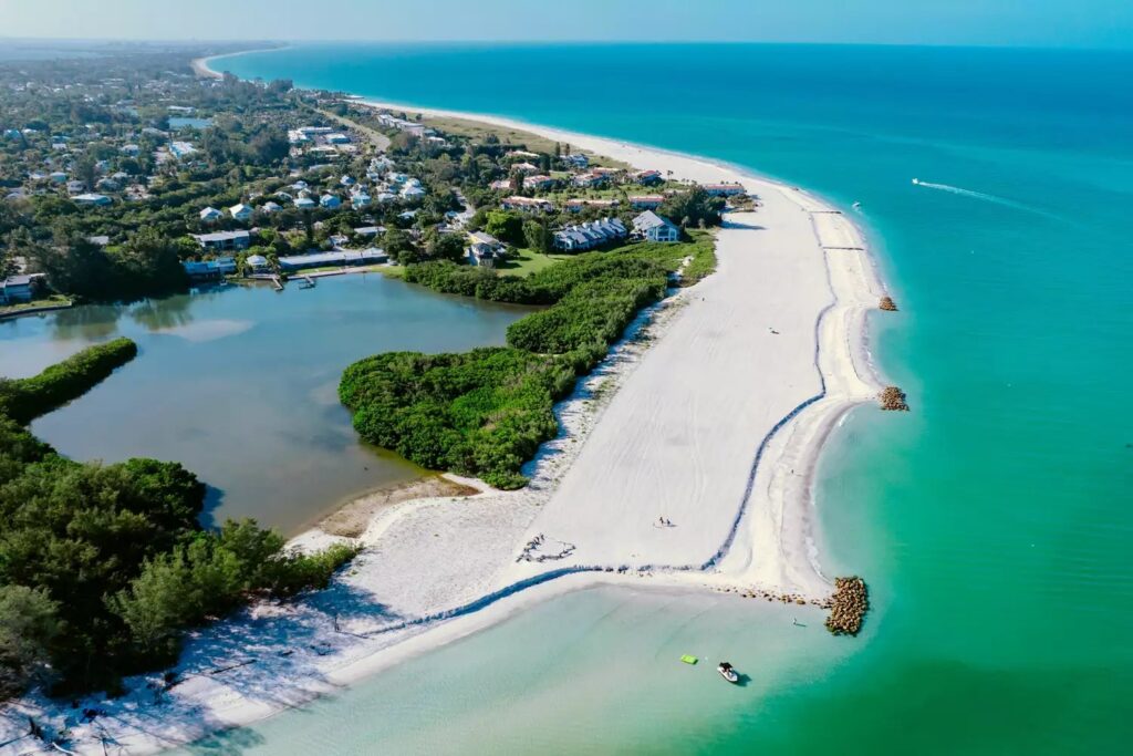 Aerial view of a sandy beach surrounded by turquoise water, with green vegetation, houses, and a small lagoon on one side, under a clear blue sky.