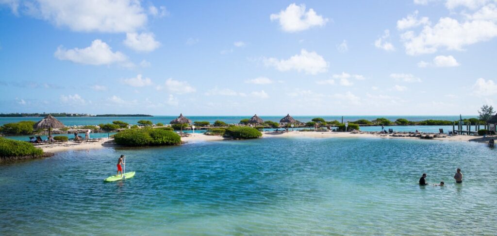 A scenic beach with clear blue water, people swimming and paddleboarding, and small thatched-roof huts along the shore under a bright sky with scattered clouds.