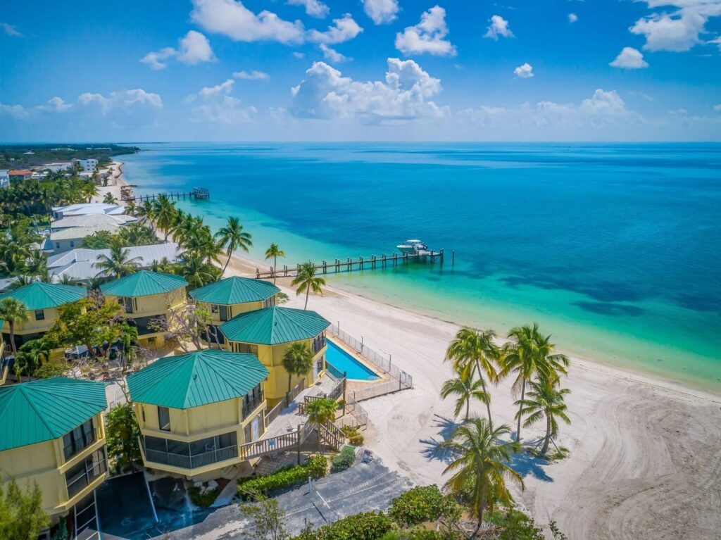 Aerial view of beachfront villas with green roofs, palm trees, white sandy beach, clear turquoise water, and a long pier stretching into the calm sea under a sunny blue sky with scattered clouds.