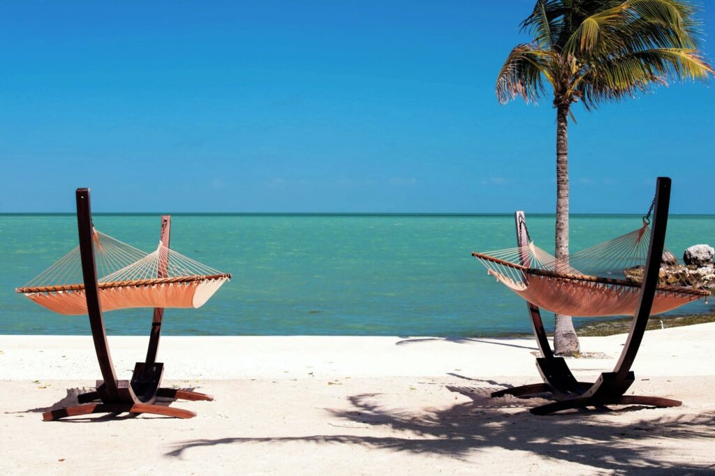 Two empty hammocks on wooden stands sit on a sandy beach facing calm turquoise water, with a palm tree nearby under a clear blue sky.