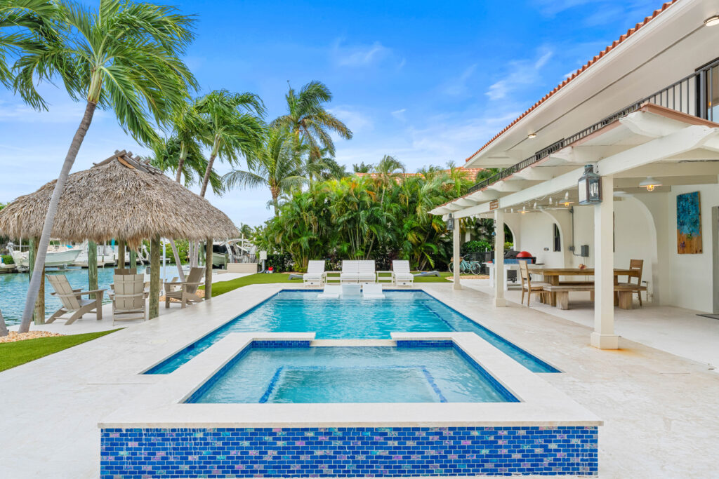 Marathon vacation rentals: deep blue. Luxury backyard with a pool and hot tub, lounge chairs, a tiki hut with seating, dining area under a pergola, and lush tropical landscaping beside a canal under a bright blue sky.