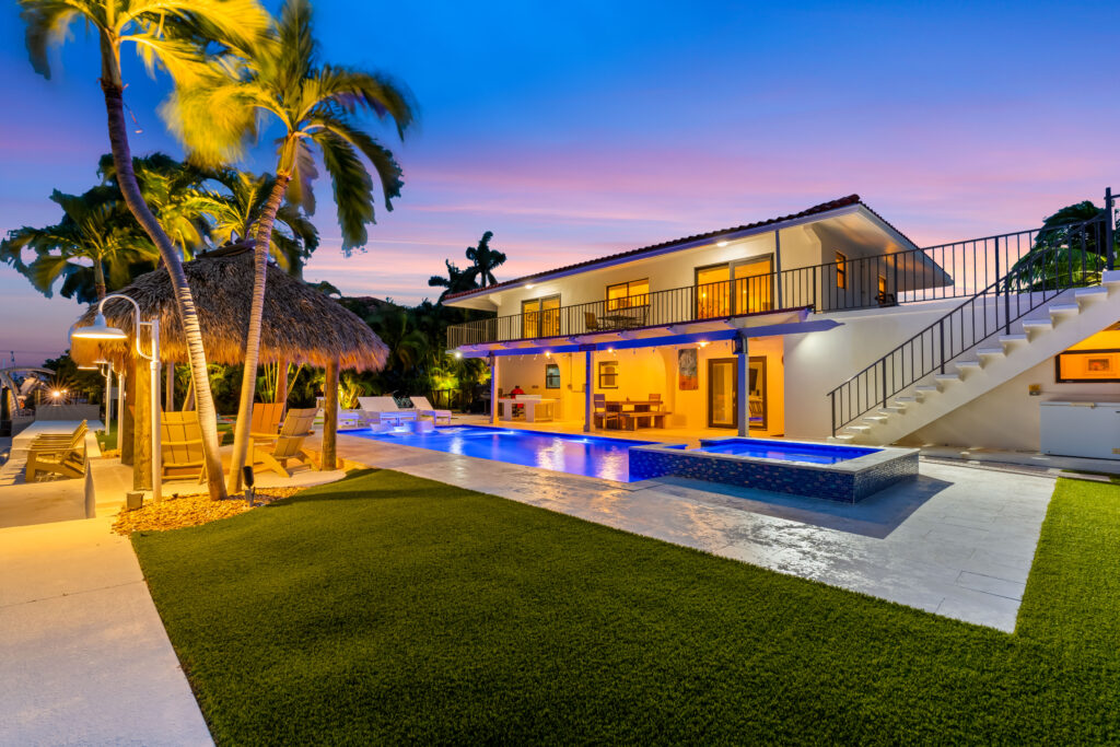 A modern two-story house with a glowing pool at sunset, surrounded by palm trees, a thatched-roof cabana, outdoor seating, and a manicured lawn under a vibrant sky—perfect for marathon vacation rentals.