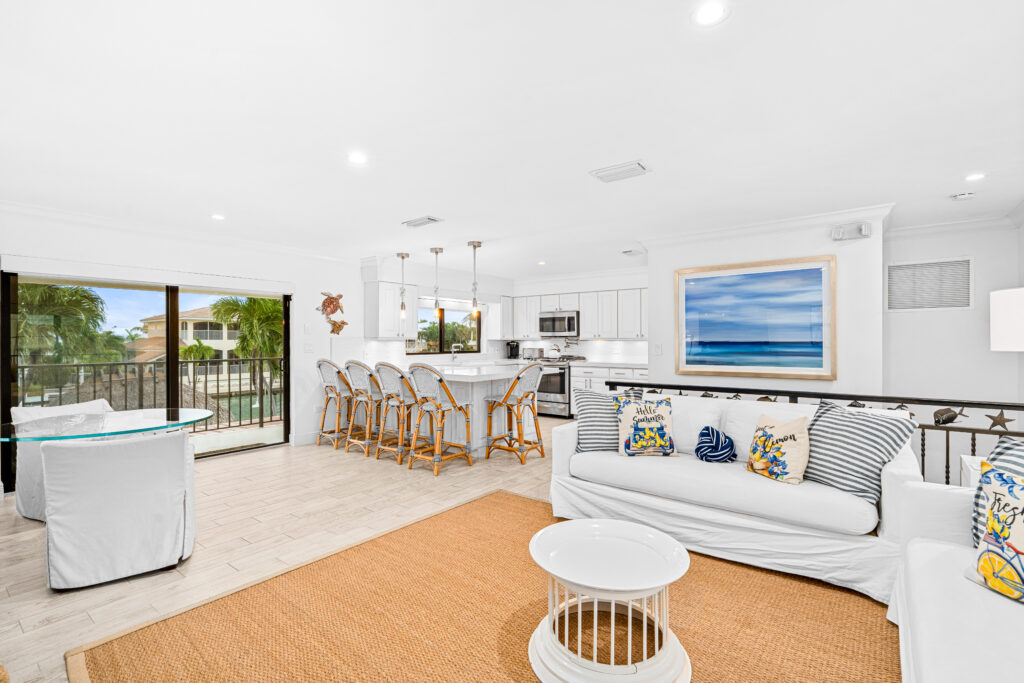 Bright, airy living room and kitchen with white furniture, blue and yellow accent pillows, barstools at a breakfast bar, large beach artwork, and sliding glass doors opening to a balcony with palm trees—perfect for marathon vacation rentals.