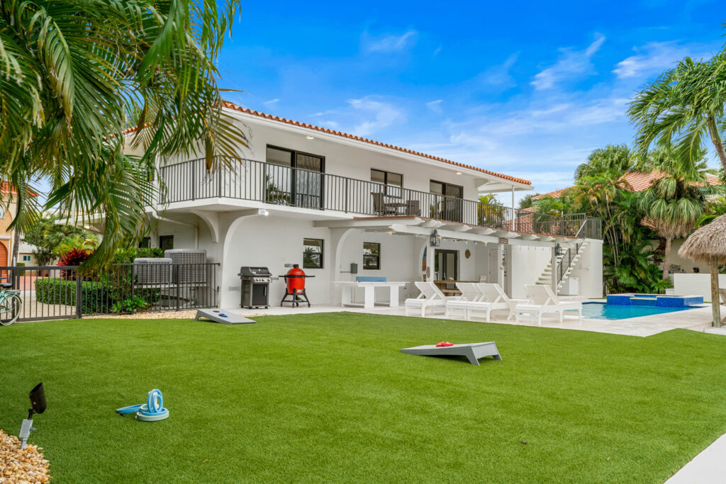A modern two-story white house with a balcony, pool, lounge chairs, grill, and outdoor games on a green lawn—one of the top marathon vacation rentals—surrounded by palm trees under a bright blue sky.