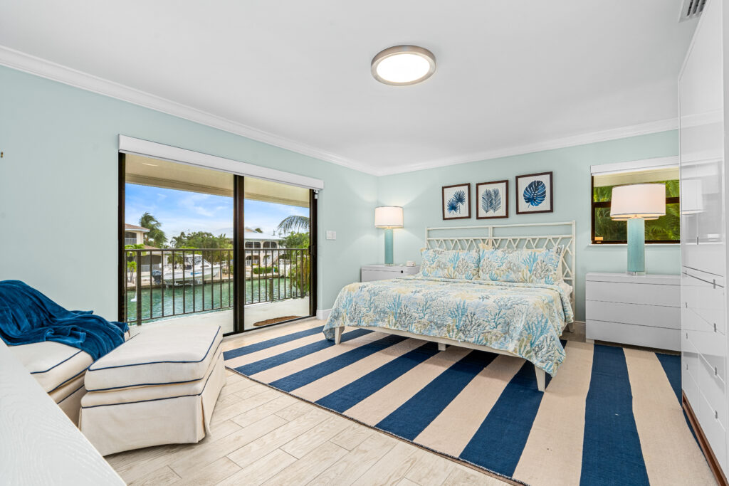 Bright bedroom with a king bed, blue and white bedding, striped rug, artwork above the bed, chaise lounge, lamp, dresser, and sliding glass doors opening to a balcony with a water view—ideal for marathon vacation rentals.