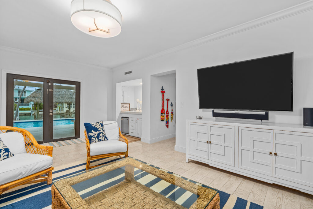 A bright living room with wicker furniture, a glass coffee table, and a large wall-mounted tv awaits in this marathon vacation rental. Sliding doors open to the pool area, showcasing coastal-themed decor with blue accents and wooden floors.