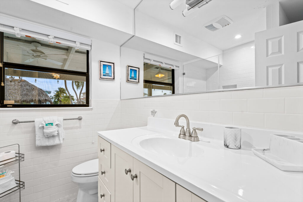 Bright, modern bathroom in one of our marathon vacation rentals features white cabinets, a wide mirror, chrome faucet, towel rack, and a window framing lush palm trees and a thatched roof. Two small framed pictures add charm to the space.