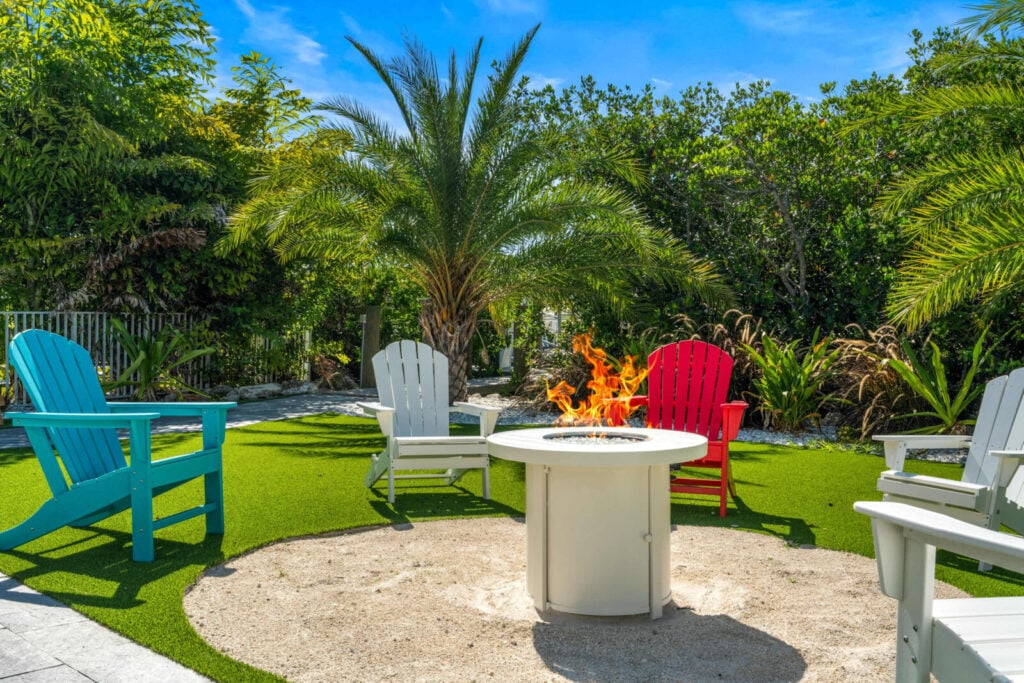Ocean muse: stunning florida keys waterfront vacation rental 5 A backyard scene with colorful adirondack chairs—blue, gray, and red—surrounding a fire pit on a sandy area, with lush green plants and palm trees in the background under a bright blue sky.