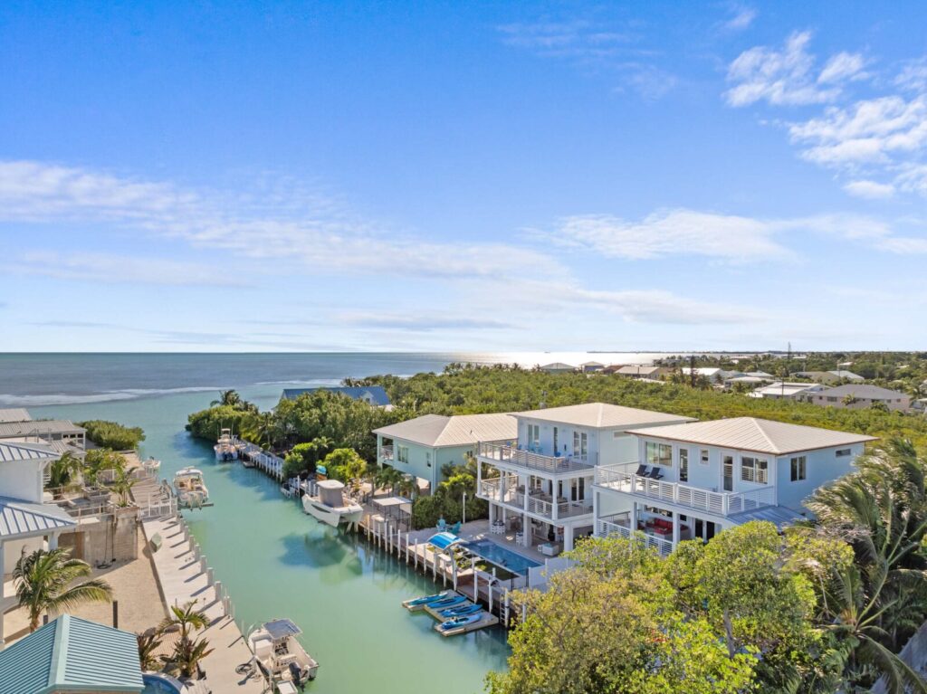 Aerial view of waterfront houses along a narrow canal with boat docks, lush greenery, and the ocean in the background—showcasing why marathon florida is perfect for snowbirds under a sunny blue sky with scattered clouds.