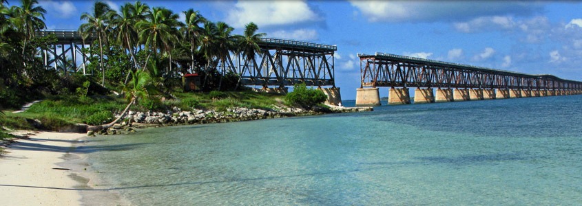 10 nature trails & parks in marathon fl | wild side guide 7 A scenic view of a sandy beach with clear turquoise water, palm trees, and an old, partially broken railway bridge stretching over the ocean under a blue sky with scattered clouds.