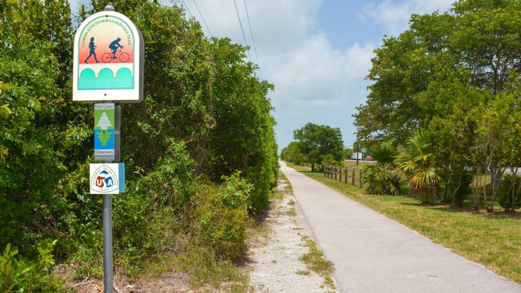 10 nature trails & parks in marathon fl | wild side guide 11 A paved walking and biking trail runs straight ahead, bordered by green trees and grass. A sign along the path shows symbols for walking and cycling. The sky is partly cloudy.