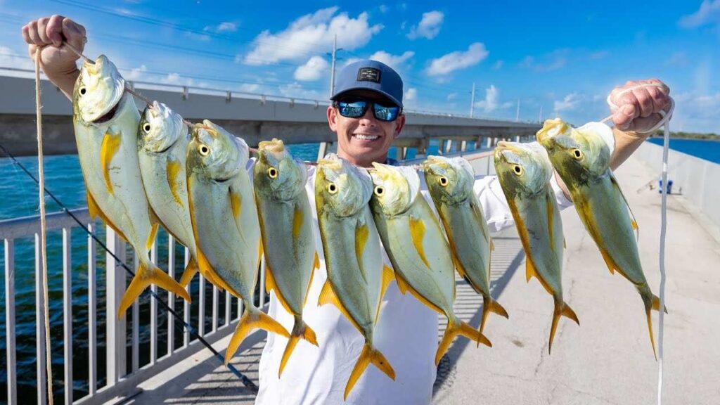 A person wearing sunglasses and a hat is smiling and holding up two lines of freshly caught yellow and silver fish on a bridge by the water under a blue sky with scattered clouds.