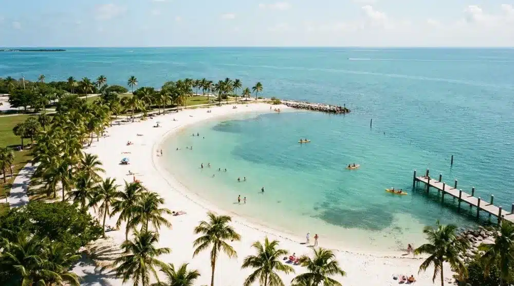 Aerial view of a tropical beach with white sand, palm trees, calm turquoise water, people swimming and sunbathing—an idyllic scene perfect for any couples getaway guide, featuring a wooden pier stretching into the ocean.