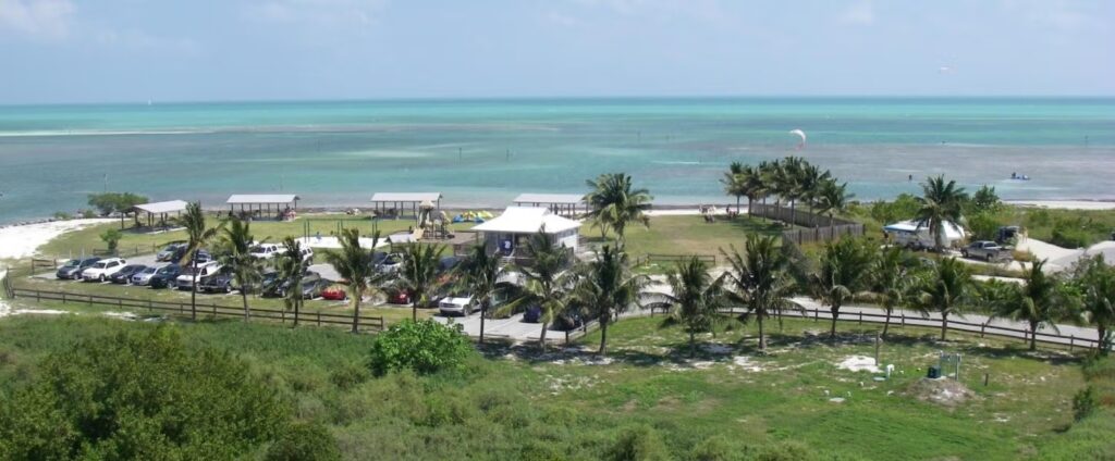 A scenic view of a beach park in marathon fl with palm trees, picnic shelters, parked cars, and a turquoise ocean under a blue sky. People can be seen enjoying outdoor activities near the shoreline—one of the top parks in marathon fl.