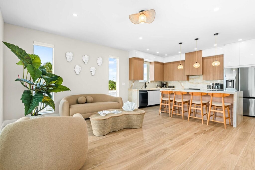 Marathon vacation rental investment: turn your home into profit 6 Modern open-concept kitchen and living room with light wood floors, beige sofa and chair, large indoor plant, and a kitchen island with four wooden stools under pendant lights. Neutral tones and natural light fill the space.
