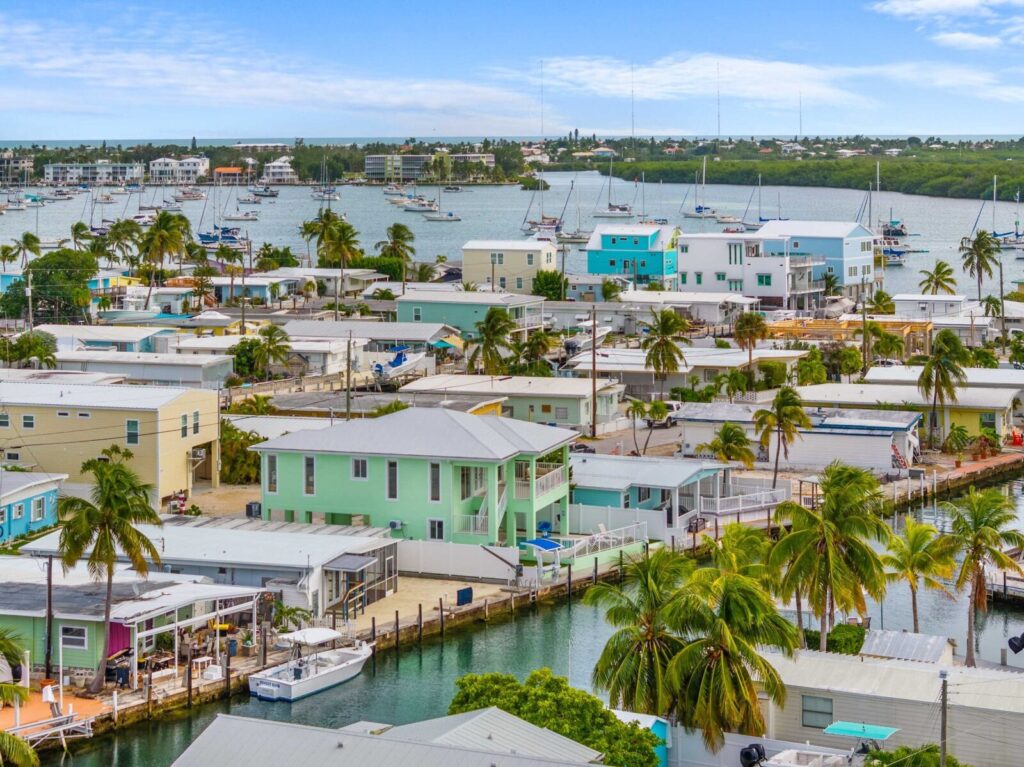 A coastal neighborhood with pastel-colored houses and palm trees lines a canal. Boats are docked along the water, and a larger harbor with sailboats is visible in the background under a partly cloudy sky.