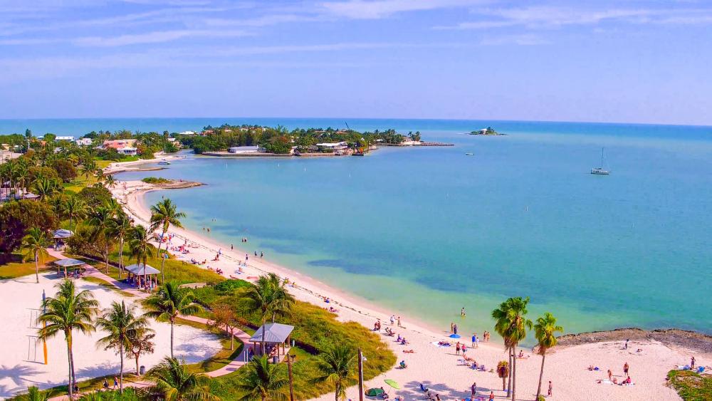 Aerial view of a tropical beach with white sand, turquoise water, palm trees, and people relaxing or swimming; small islands and sailboats are visible in the calm ocean background under a blue sky.