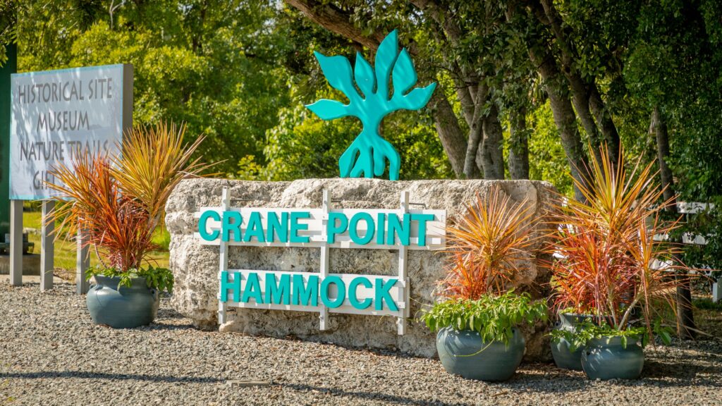 A stone sign reading crane point hammock in teal letters stands surrounded by potted plants, with trees and a secondary sign for a historical site, museum, and nature trails in the background.