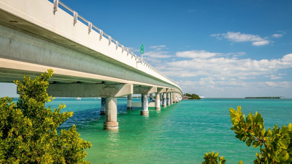 A long concrete bridge stretches over turquoise water, supported by pillars, with lush green foliage in the foreground and small islands in the distance under a blue sky with scattered clouds.