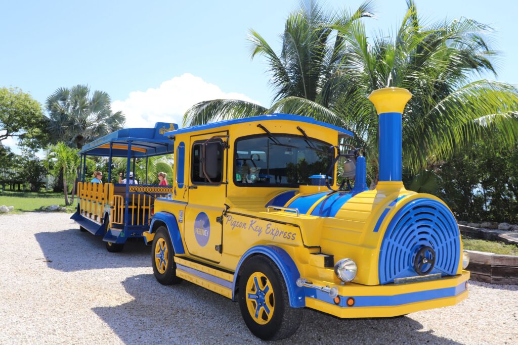 A bright yellow and blue open-air tour train labeled key express is parked on a gravel path, with palm trees and green foliage in the background under a sunny sky.