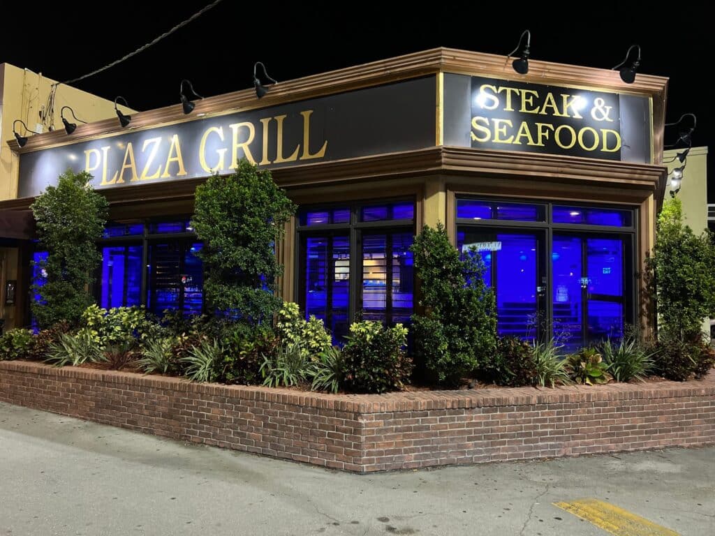 Nighttime view of plaza grill restaurant with a large black and gold steak & seafood sign. Blue lighting glows from inside, and the exterior is lined with bushes—an inviting spot for any couples getaway guide.