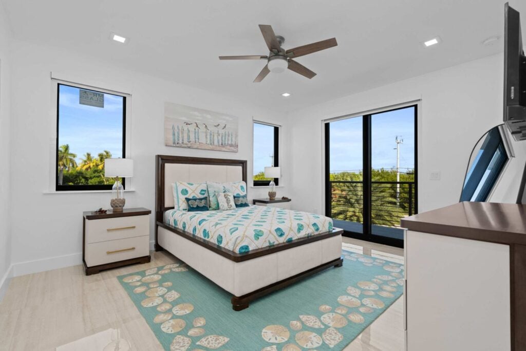 Ocean muse: stunning florida keys waterfront vacation rental 6 A modern, bright bedroom with large windows, a ceiling fan, a bed with patterned teal and beige bedding, matching nightstands and lamps, a dresser, and a blue area rug with shell designs. Trees are visible outside.