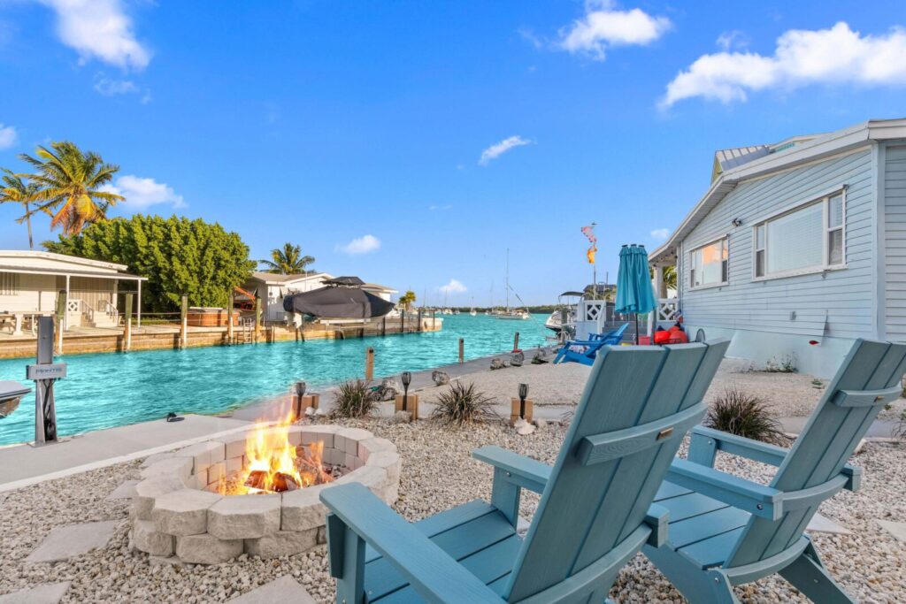 9 romantic anniversary ideas in marathon, fl | couples getaway guide 2 Two blue adirondack chairs by a stone firepit overlook a turquoise canal with boats docked nearby, surrounded by gravel and tropical plants beside a light-blue waterfront house under a clear, sunny sky.