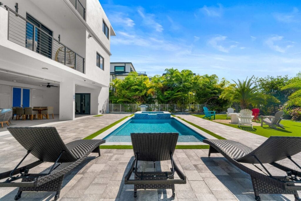 Ocean muse: stunning florida keys waterfront vacation rental 7 Modern backyard with a rectangular pool, lounge chairs, jacuzzi, patio seating, and colorful adirondack chairs surrounded by lush greenery and a contemporary white house under a blue sky.