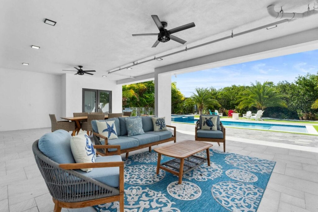 Ocean muse: stunning florida keys waterfront vacation rental 8 Covered patio with ceiling fans, outdoor sofa set, and a blue patterned rug. Beyond the patio is a swimming pool surrounded by greenery, with lounge chairs and clear blue sky in the background.