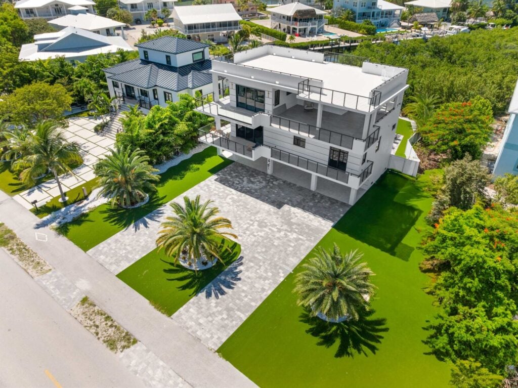 Ocean muse: stunning florida keys waterfront vacation rental 12 A modern white three-story house with balconies and a paved driveway, surrounded by a manicured lawn, palm trees, and neighboring homes under a clear sky.