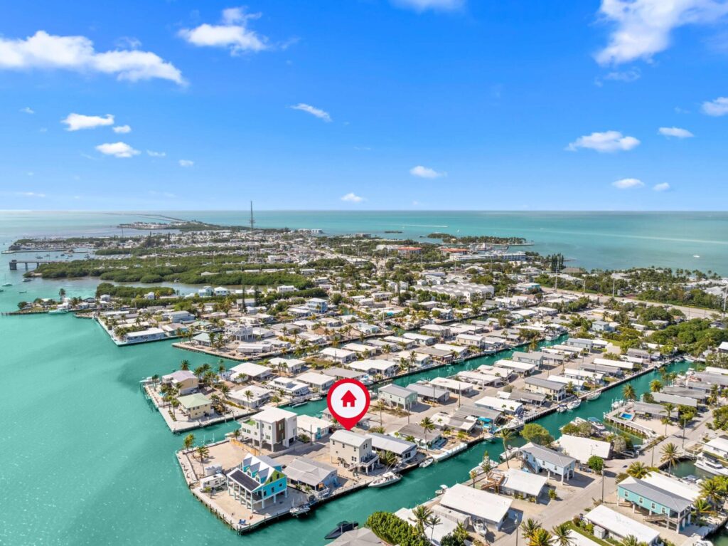 Marathon vacation rental investment: turn your home into profit 2 Aerial view of a coastal neighborhood with houses along turquoise canals, bordered by the ocean. A red marker with a house icon highlights one property. The sky is clear with a few clouds.