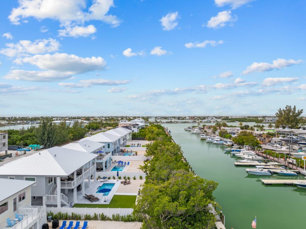 9 romantic anniversary ideas in marathon, fl | couples getaway guide 6 Aerial view of waterfront homes with white roofs along a calm canal lined with boats on a sunny day, featuring green trees, blue lounge chairs by pools, and a bright blue sky with scattered clouds.