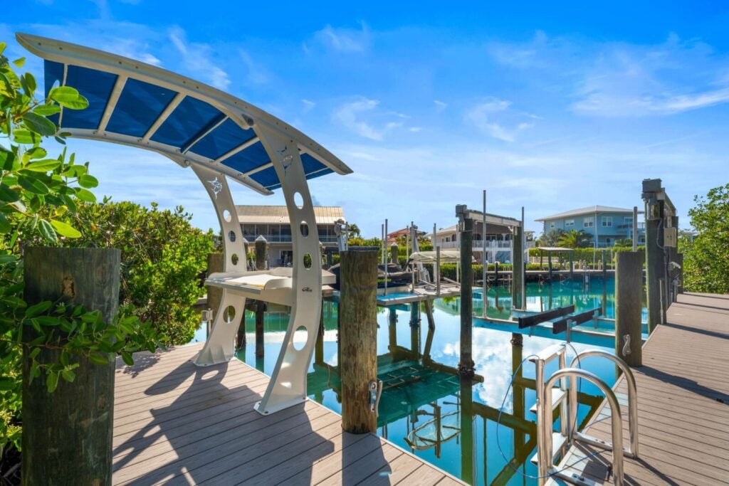 Ocean muse: stunning florida keys waterfront vacation rental 9 A modern wooden dock with a curved, blue-and-white shaded bench overlooks a calm, reflective canal lined with houses and boats under a bright blue sky. Greenery borders the dock, and a swim ladder descends into the water.