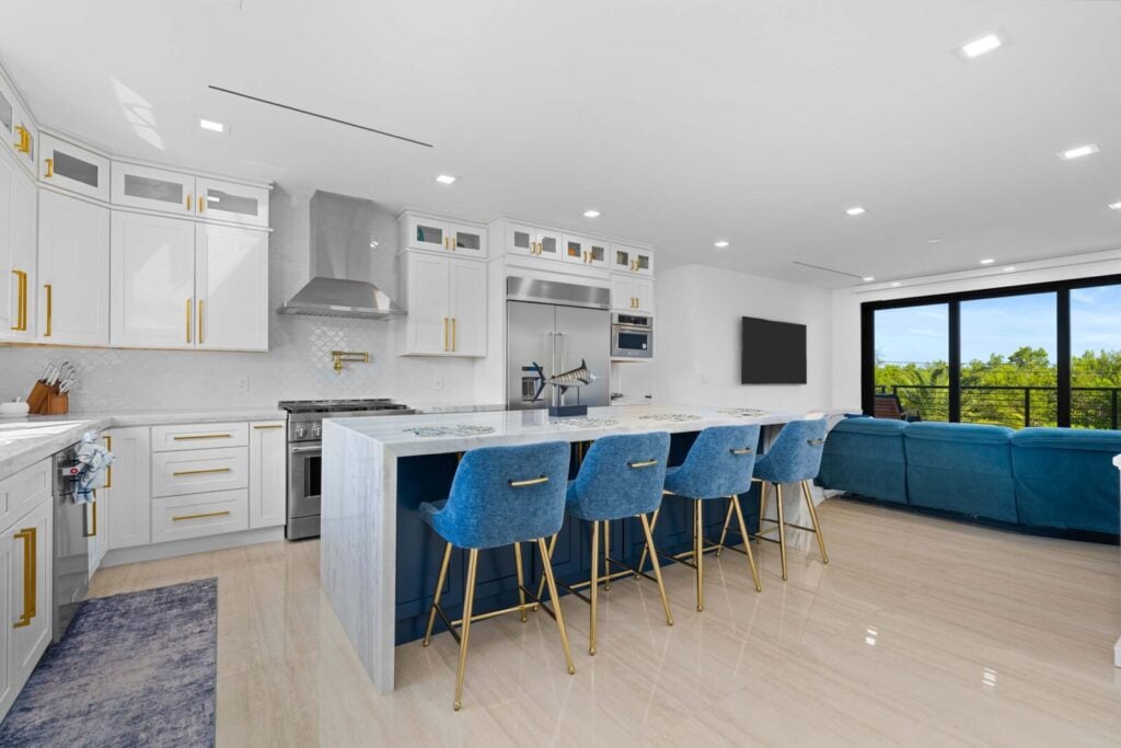 Ocean muse: stunning florida keys waterfront vacation rental 4 Modern kitchen with white cabinets, gold handles, stainless steel appliances, and a large island featuring four blue barstools. Open floor plan leads to a living area with a blue sofa and large windows overlooking greenery.
