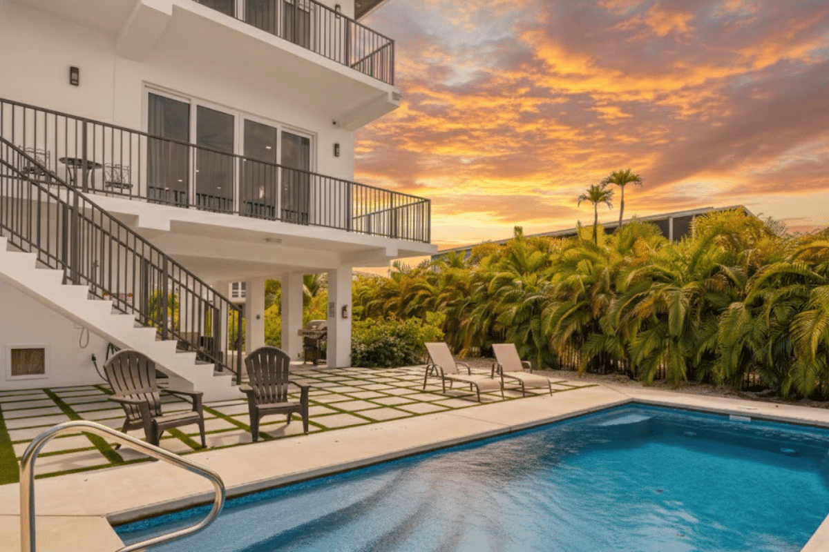 Where to watch the sunset in marathon fl | 7 stunning spots 2 Modern house with balconies overlooks a pool surrounded by lounge chairs and palm trees, set under a vibrant orange sunset sky.