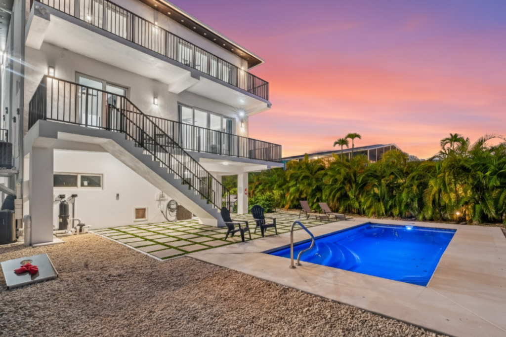 Where to watch the sunset in marathon fl | 7 stunning spots 6 Modern two-story house with balconies, an outdoor staircase, a lit swimming pool, lounge chairs, and lush palm trees at sunset. The sky is vibrant with shades of orange, purple, and pink.
