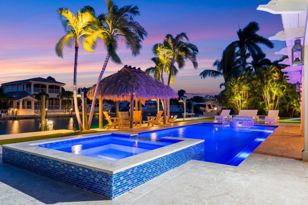 10 upgrades to increase your florida keys vacation rental revenue (2026) 3 A luxurious pool and hot tub illuminated at dusk, surrounded by palm trees, lounge chairs, a thatched tiki hut, and waterfront homes, with a colorful sunset sky in the background.