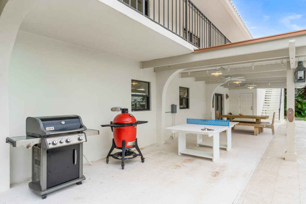 10 upgrades to increase your florida keys vacation rental revenue (2026) 6 Covered outdoor patio with a black gas grill, a red ceramic smoker, a blue and white ping-pong table, and a wooden dining table with benches; white walls and overhead lights create a bright, welcoming space.