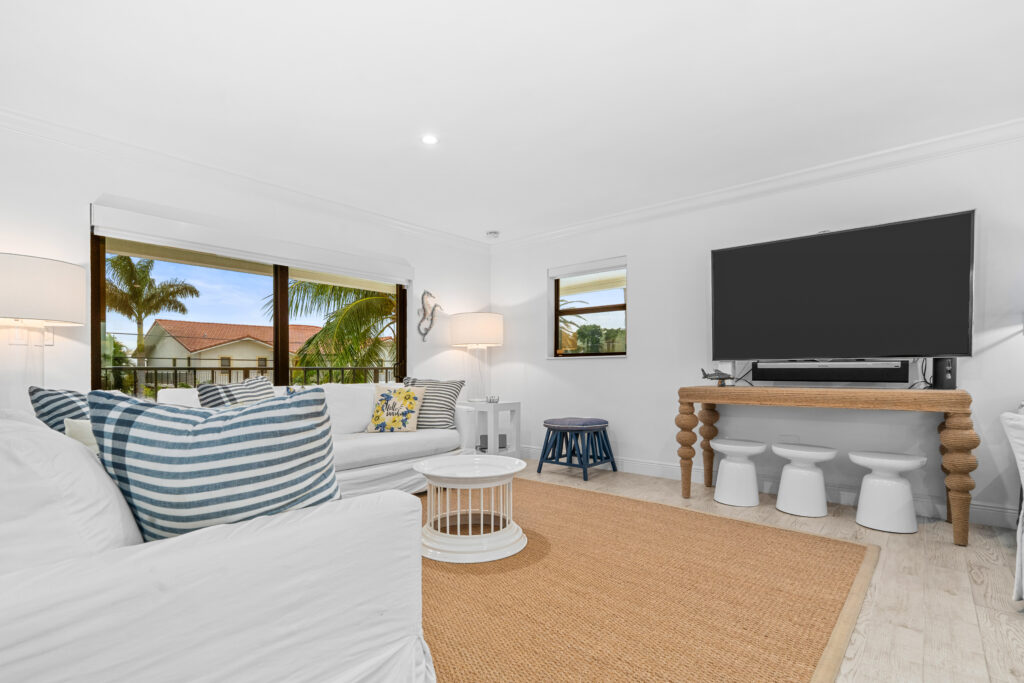 10 upgrades to increase your florida keys vacation rental revenue (2026) 9 Bright living room with white sofas, striped pillows, a round coffee table, large tv on a wooden console, woven rug, and windows showing palm trees and a building outside.