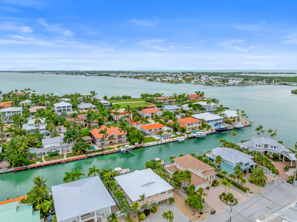 10 upgrades to increase your florida keys vacation rental revenue (2026) 10 Aerial view of waterfront homes with docks and boats along a turquoise canal, bordered by palm trees, near a large bay under a blue sky with scattered clouds.