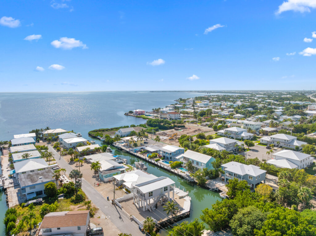 Aerial view of a coastal neighborhood with white-roofed houses, a canal running between homes, palm trees, and calm blue ocean under a clear sky with a few scattered clouds.