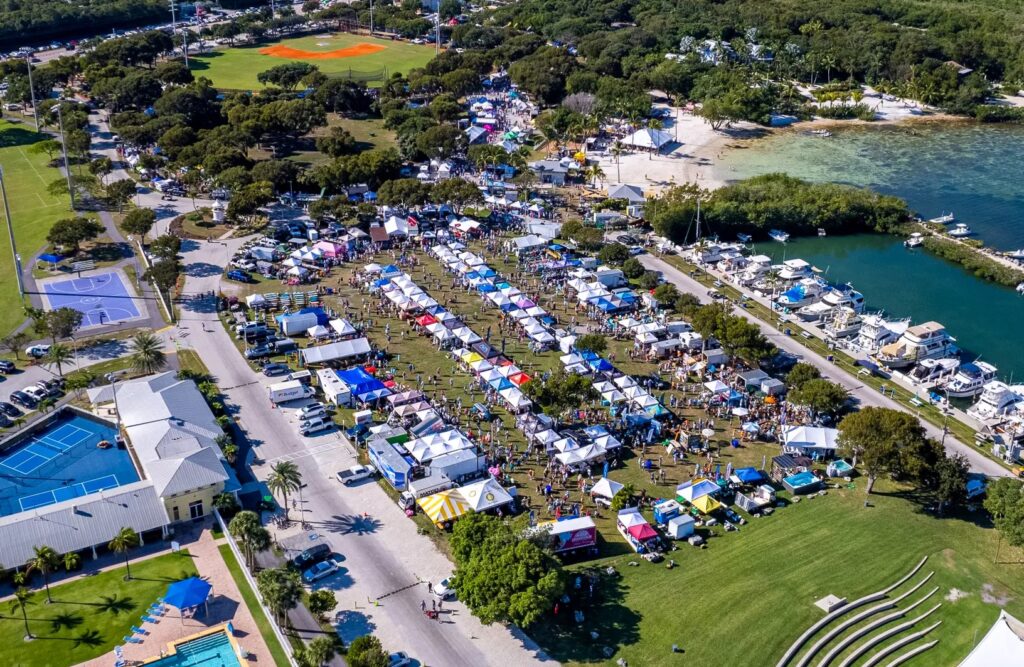 Aerial view of an outdoor festival with rows of colorful tents, crowds of people, food trucks, nearby boats at a marina, sports courts, green lawns, and trees, set next to a waterfront on a sunny day.