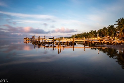 Where to watch the sunset in marathon fl | 7 stunning spots 5 A wooden pier extends into calm, reflective water at sunrise or sunset, with palm trees and a sandy shoreline in the background under a partly cloudy sky.