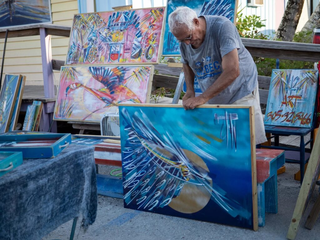 An older man with gray hair and glasses is arranging colorful abstract paintings at an outdoor art display, with multiple vibrant artworks leaning against a wooden platform behind him.