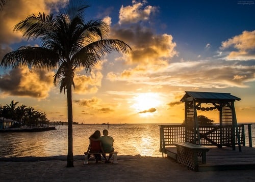 Where to watch the sunset in marathon fl | 7 stunning spots 3 A couple sits on a bench under a palm tree, watching a vibrant sunset over a calm waterfront. Silhouetted structures and clouds frame the golden sky, creating a peaceful, romantic atmosphere.